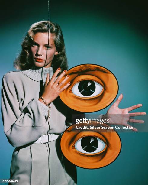 American actress Lauren Bacall poses behind a pane of glass decorated with two giant eyes, circa 1950.