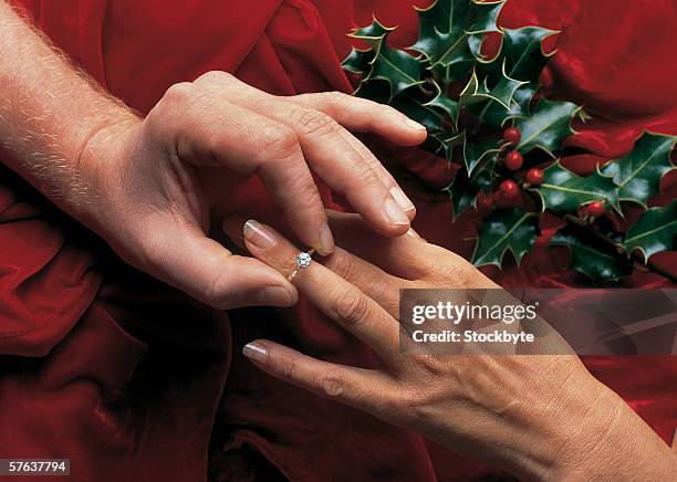 close-up of a man's hand putting a diamond ring on a woman's finger beside a sprig of holly - woman putting on diamond stock pictures, royalty-free photos & images