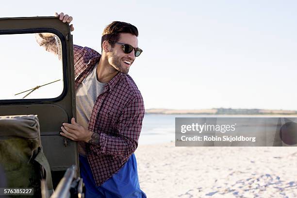 young man standing by old jeep on beach - auto stehend stock-fotos und bilder