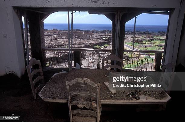 Dinning table covered under ash with a view of Plymouth and the Caribbean Sea in background. 10 years ago the Soufriere-Hills Volcano erupted and...