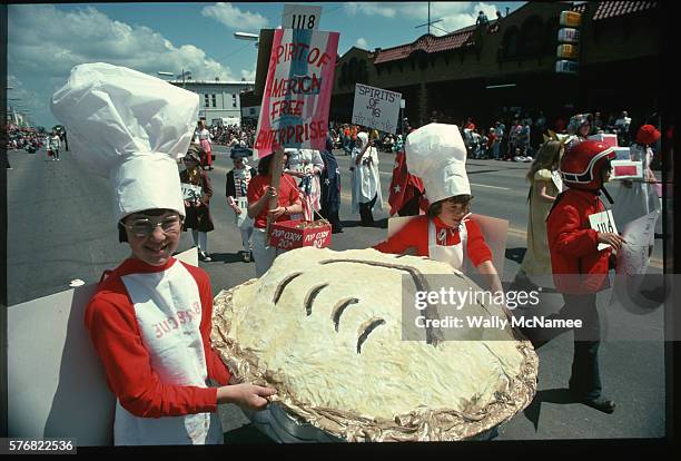 Children in a Bicentennial parade push a large paper-mache apple pie in celebration of American independence. Behind them other children are dressed...