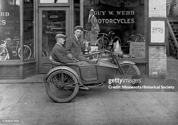 In front of Guy Webb's Minneapolis motorcycle shop, two men ride a Harley-Davidson motorcycle equipped with a side car.