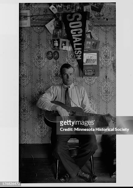 Man sits below a display of photographs and a Socialist pennant, playing a guitar.