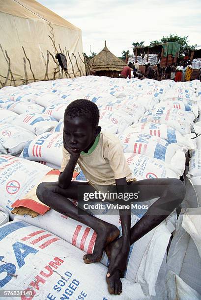 Young Dinka boy sits on sacks of sorghum sent as aid from the United States to the refugee camp of Atepe, Sudan. Civil war and widespread famine have...