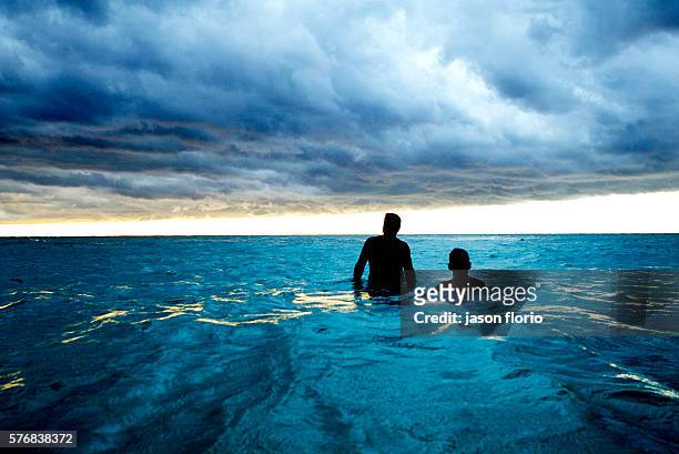Two silhouetted men with their backs to the camera stand in the sea watching storm clouds gather overhead at Playa Del Este, Cuba.