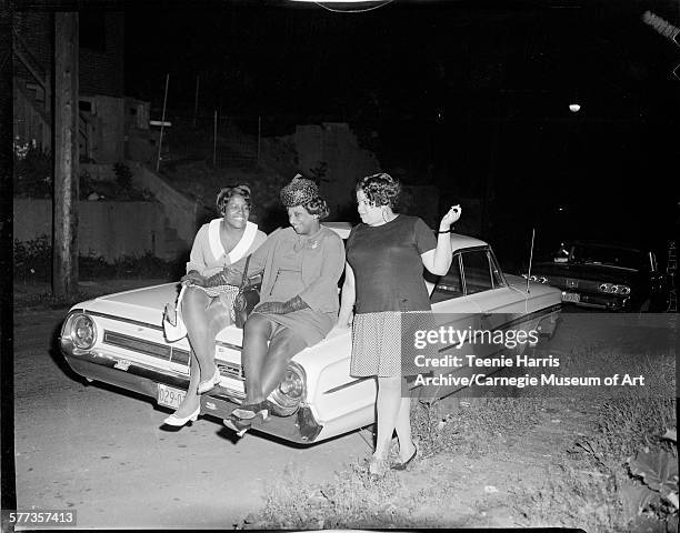Three women, including two seated on trunk of light colored Ford Galaxie car, with Pennsylvania license plate reading '029-0', parked on side of...