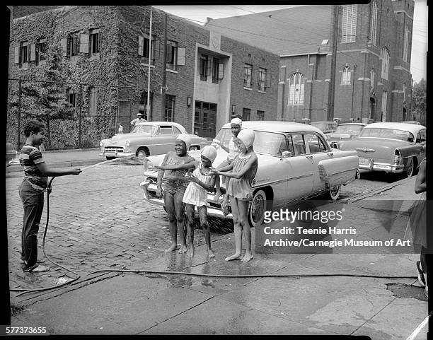 Boy wearing striped t-shirt aiming hose at four girls wearing bathing caps and swimming or sun suits, on sidewalk and block street, with parked cars,...