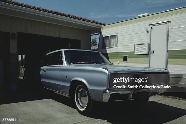 Car and caravan camper parked in the driveway of a suburban home, the car emerging from the garage, half in shadow, 1966.