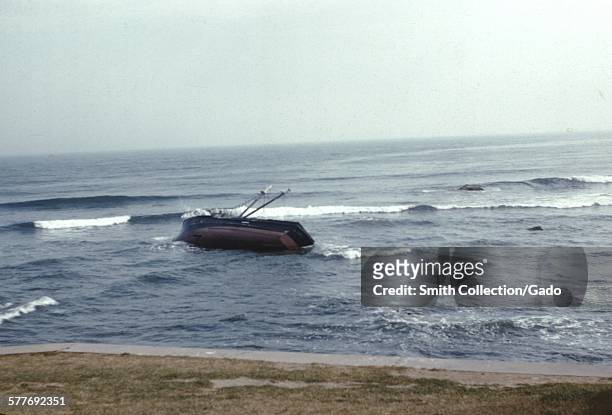 Fishing boat foundering and sinking in a storm, rolled onto its side as swells of waves push it into shore, Gloucester, Massachusetts, 1952.