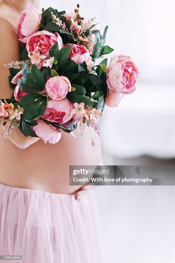 Hands and pink peony flowers on pregnant belly of a woman in a pink tender tutu skirt