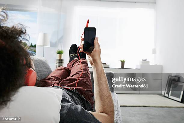 young man lying on couch at home listening music with headphones and smartphone - back of sofa stock pictures, royalty-free photos & images