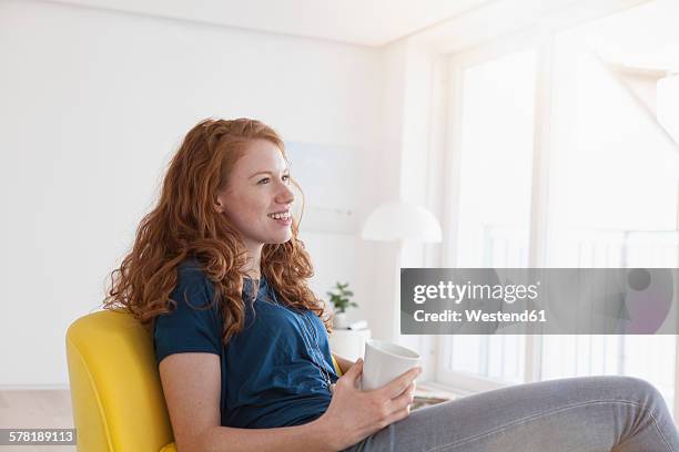 happy young woman with cup of coffee sitting in her living room - donna poltrona foto e immagini stock