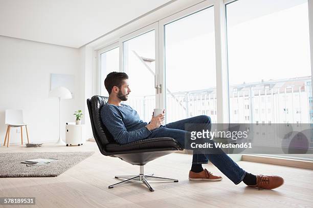 young man relaxing on leather chair with cup of coffee in his living room - coffee window stock pictures, royalty-free photos & images