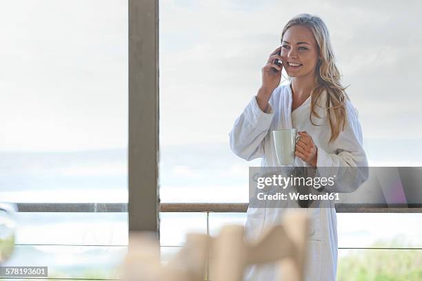 young woman wearing bathrobe talking on cell phone in beach house - beach house balcony fotografías e imágenes de stock