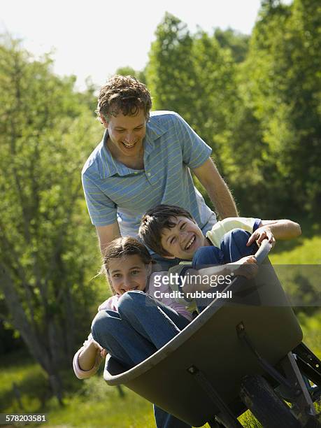 man pushing his son and daughter in a wheelbarrow - camiseta stock pictures, royalty-free photos & images