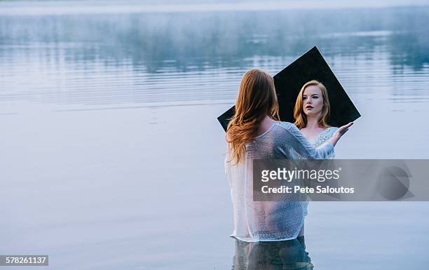 rear view of young woman standing in lake holding mirror looking at reflection - schneewittchen stock-fotos und bilder