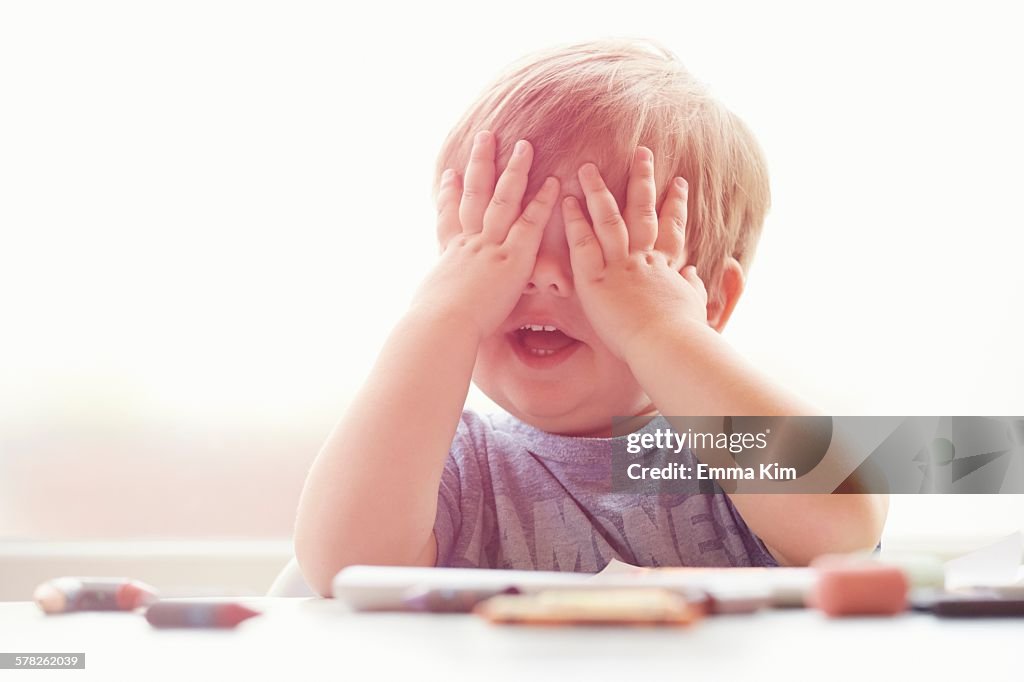 Boy sitting at table resting on elbows, mouth open, covering eyes with hands