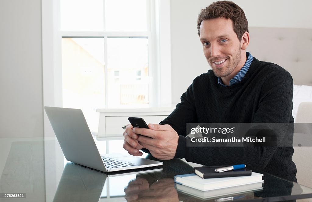 Mid adult man sitting at table, using laptop and smartphone