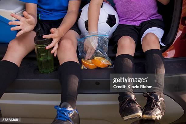 waist down of boy and younger sister sitting in car boot eating oranges on football practice break - sports car photos et images de collection