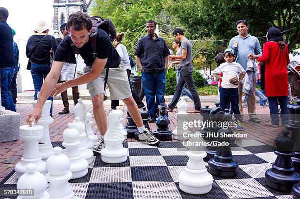 Man wearing a backpack reaches to move the king piece from a life-sized chess set, while onlookers behind him observe his strategy, during Baltimore...