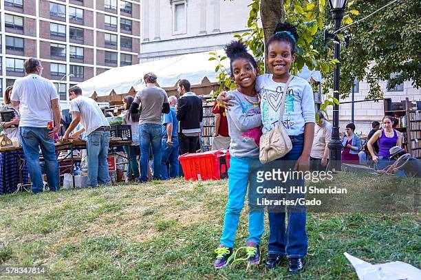 Two young sisters stand with arms around each other while they pose for their photo, with a tree and several tables and tents of used books for sale...