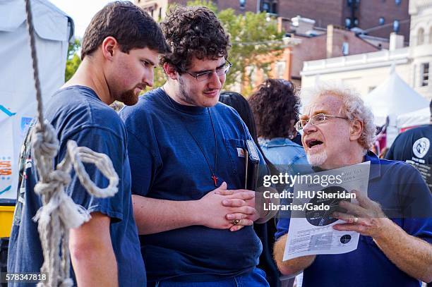 An older man with glasses reads from a flyer about income taxes to two young men wearing crosses and holding bibles, during Baltimore Book Festival,...