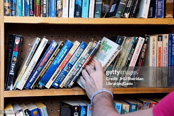 Man's arm reaches for a used paperback book from a shelf of leaning books for sale during Baltimore Book Festival, Baltimore, Maryland, September,...