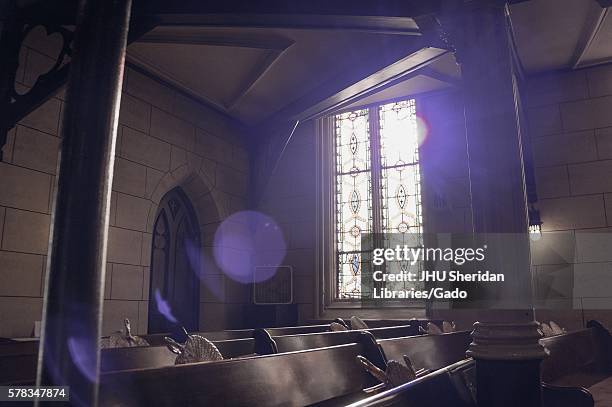 Sun filters through the stained glass of an old church, creating shadows on the pews with woven fans in their holders, Baltimore, Maryland,...