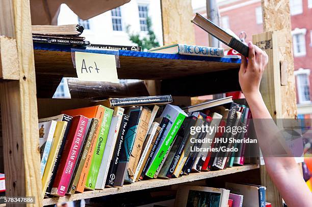 Hand reaches for a book on the top shelf of a cart labeled "Art, " with used books for sale during Baltimore Book Festival, Baltimore, Maryland,...