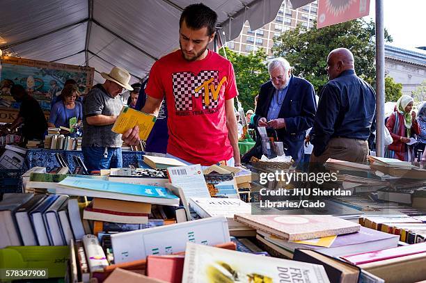 Young man wearing a MTV t-shirt exploring a large pile of books among other people exploring books at a tent at the Baltimore Book Festival,...