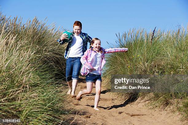 girl and boy running through marram grass holding football smiling - shorts down stock pictures, royalty-free photos & images