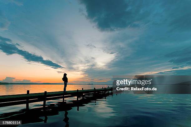 silhouette of woman on lakeside jetty with majestic sunset cloudscape - scenics stock pictures, royalty-free photos & images