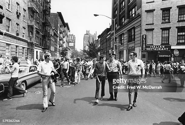 One month after the demonstrations and conflict at the Stonewall Inn, activist Marty Robinson , a police officer, and others walk in the street with...
