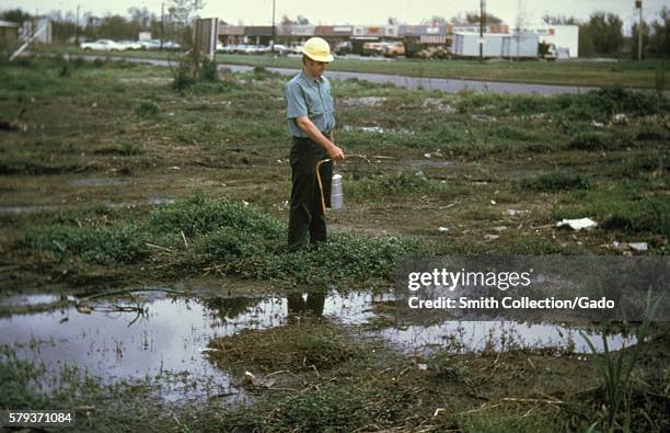 This photograph shows a man using a hand-carried compressed air sprayer apply larviciding oil to mosquito breeding pools, 1981. Standing pools of...