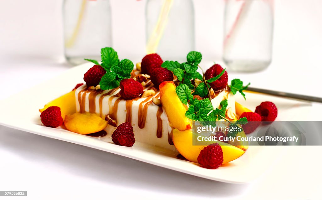 Home made vanilla ice cream with caramel sauce, hazelnuts, peach, raspberries and mint on the rectangular plate. Bottles of lemonade are on the background. On white.