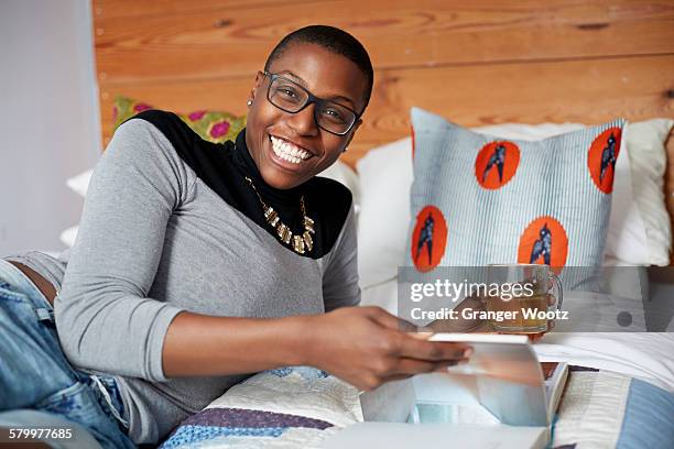 mixed race woman drinking tea and reading on bed - mixed race woman drinking tea stock-fotos und bilder