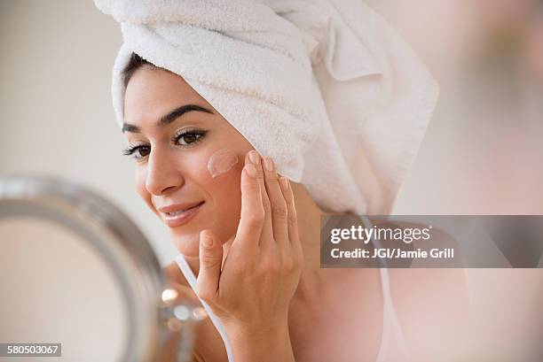 woman with hair in towel rubbing lotion on face - soin de la peau photos et images de collection