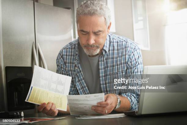 hispanic man paying bills on laptop in kitchen - bills payment stock-fotos und bilder