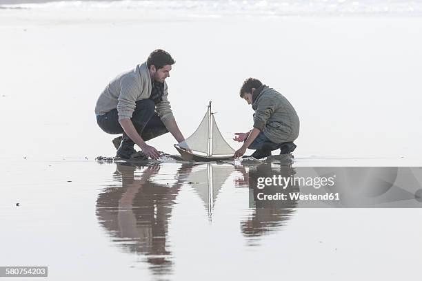 father and son playing with toy sailing boat at seafront - father son sailing stock-fotos und bilder