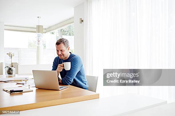 man working at home using laptop drinking coffee - working at home desktop computer stock-fotos und bilder