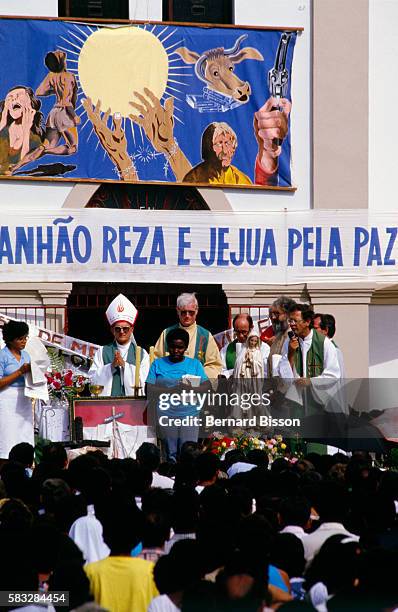 Priest Davi Alves da Silva preaches from a raised platform outside a church to a crowd of followers of Liberation Theology. The promotion of direct...