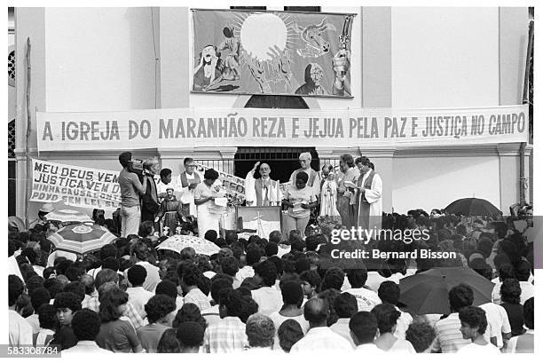 Densely packed crowd of followers attends a Liberation Theology service, given from a raised platform outside a village church decorated with...