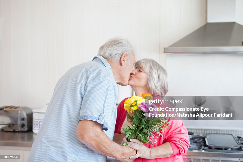 Woman kissing senior man with flowers in kitchen