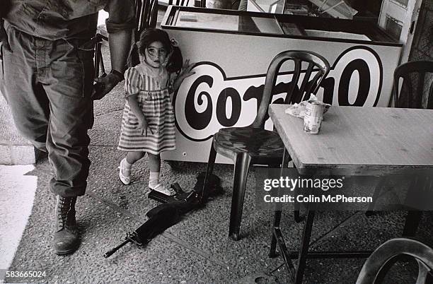 Young settler girl pointing to a gun belonging to a soldier from the Israel Defense Force at a cafe in the West Bank settlement of Kiryat Arba near...