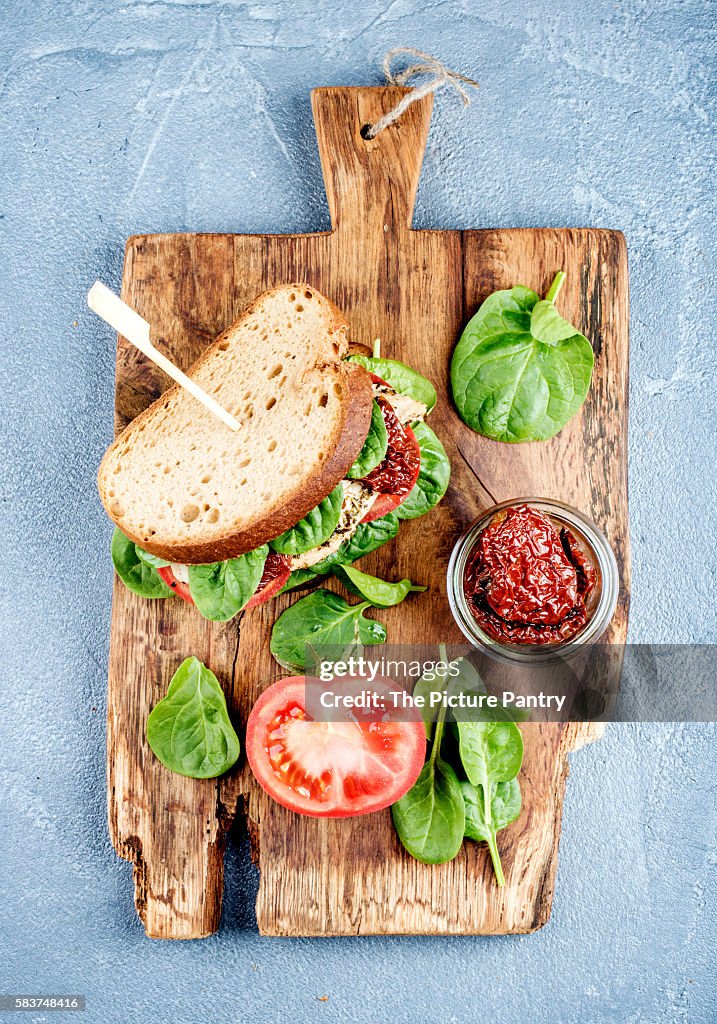 Chicken, sun-dried tomato and spinach sandwich with spicy sauce on rustic wooden board over grey concrete textured background