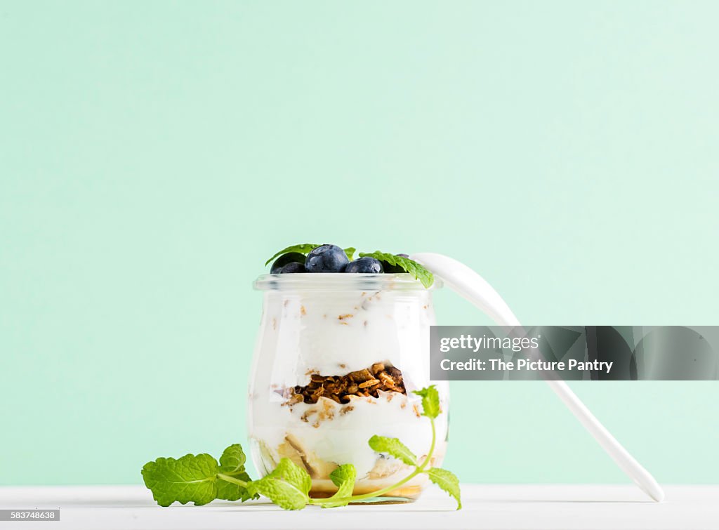 Yogurt oat granola with fresh berries and mint leaves in glass jar on grey concrete textured backdrop