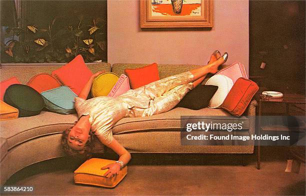 Vintage color photograph of a woman infold satin pajamas reeling upside down on a sectional sofa full of brightly colored pillows, 1950s.