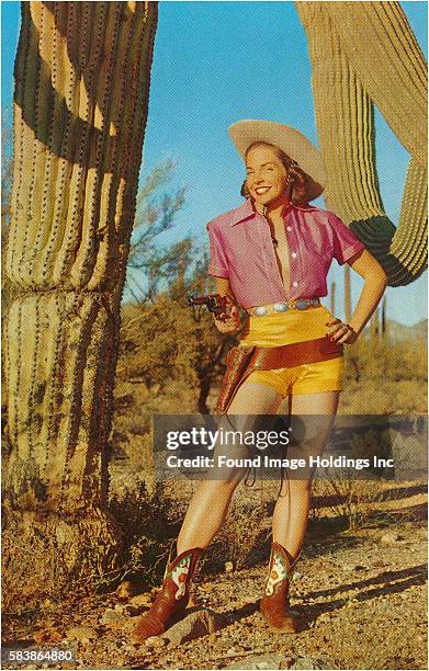 Vintage color photograph of a faux cowgirl ,standing amidst cacti in the desert, wearing yellow satin short shorts, an unbuttoned pink blouse, a...