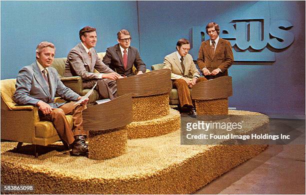 Vintage color photograph of five newscasters in suits and ties on a carpeted platform set, 1960s. The word ‘News” is in large, raised, blue letters...