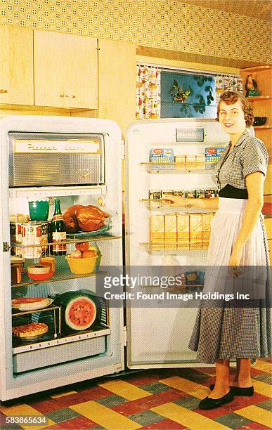 Vintage color photograph of a kitchen interior in which a housewife wearing a dress with a white apron stands in front of a fully loaded open...
